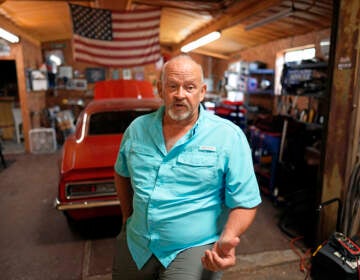 A man stands, speaking, in the foreground of the photo, with a red car and the American flag visible behind him.