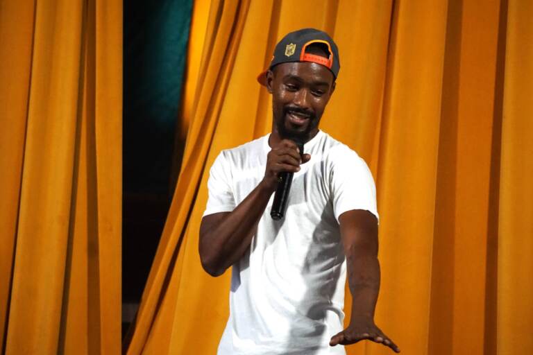 A person holds a microphone and gestures. Stage curtains are visible in the background.
